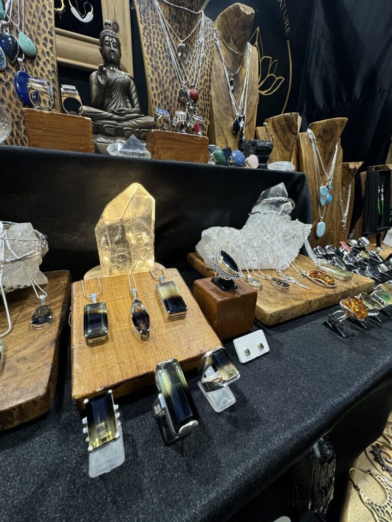 A close view of the Elements by Nature stall table at Old Spitalfields Market, smoky quartz and amber pendants and silver rings laid on wooden boards beside a lit quartz point.