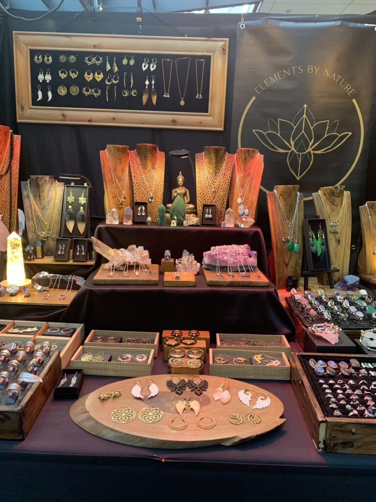 The Elements by Nature stall at Old Spitalfields Market, seen head on: a framed board of earrings, wooden busts hung with gemstone pendants, crystal points and trays of silver rings beneath the gold lotus banner.